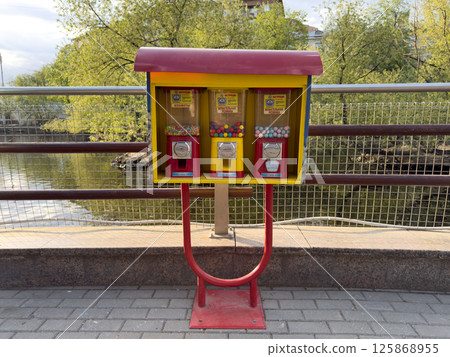 Colorful candy and gum vending machine stands on paved riverside path, inviting passersby to enjoy sweet treat. Surrounded by greenery, adds playful touch to the urban scene Colorful candy and gum vending machine stands on paved riverside path, inviting passersby to enjoy sweet treat. Surrounded by greenery, adds playful touch to the urban scene 125868955
