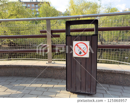 Urban trash bin with clear no smoking sign, located in clean public area near water and greenery, symbolizing smoke free lifestyle, public health awareness and city regulations Urban trash bin with clear no smoking sign, located in clean public area near water and greenery, symbolizing smoke free lifestyle, public health awareness and city regulations 125868957