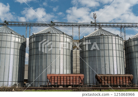 Three metallic grain silos stand tall, surrounded by a scenic countryside, under an expansive blue sky dotted with clouds 125868966