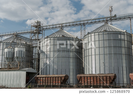 Large metallic silos feature prominently at a grain storage facility amidst a cloudy sky and scattered fields Large metallic silos feature prominently at a grain storage facility amidst a cloudy sky and scattered fields 125868970