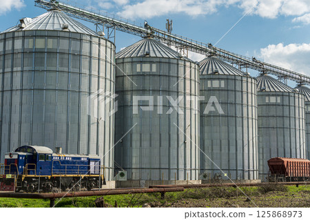A blue locomotive rests beside a row of towering metal silos in a rural area, showcasing industrial agriculture and machinery A blue locomotive rests beside a row of towering metal silos in a rural area, showcasing industrial agriculture and machinery 125868973