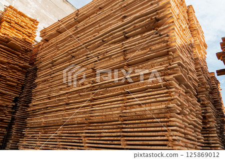 Wood planks piled high in a lumber yard, showcasing organized stacks under blue skies during the day 125869012