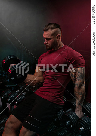 muscular man in a red shirt is lifting a dumbbell in a fitness studio. He appears focused and determined while surrounded by various weights. The lighting creates an intense atmosphere. 125870090