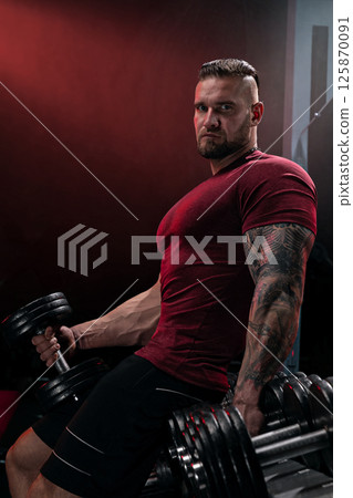 muscular man engages in strength training by lifting heavy dumbbells in a dimly lit gym. The ambiance creates a focused atmosphere for workouts. 125870091