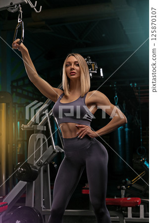 fitness enthusiast demonstrates strength training using a cable machine in a dimly lit gym space. The focus is on posture and muscle definition highlighting fitness commitment. 125870107