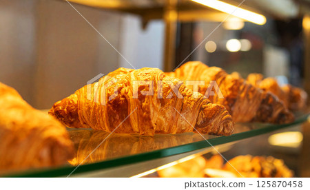 golden croissant, buttery pastry, croissants arranged on a shelf in a bakery display case, The warm, golden lighting illuminates the flaky texture, deliciousness and appeal of freshly baked goods 125870458