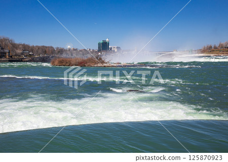 Visitors admire the breathtaking views of Niagara Falls under clear skies, as the majestic waterfall cascades powerfully into the river. 125870923