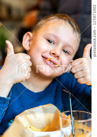Smiling boy showing thumbs up with fries in mouth, concept of childhood joy and fun eating Smiling boy showing thumbs up with fries in mouth, concept of childhood joy and fun eating 125871002