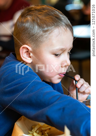 Young boy eating French fries and sipping a drink through a straw at a restaurant table close-up view, concept of casual dining Young boy eating French fries and sipping a drink through a straw at a restaurant table close-up view, concept of casual dining 125871016