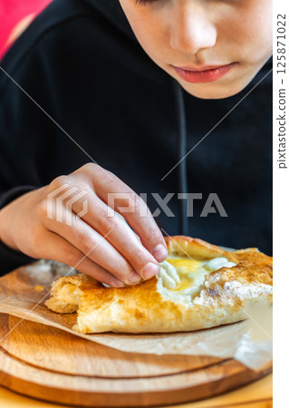 Boy eating traditional khachapuri bread with cheese and egg filling on wooden board close-up view, concept of traditional cuisine enjoyment 125871022