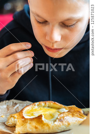 Boy eating traditional khachapuri bread with cheese and egg filling on wooden board close-up view, concept of traditional cuisine enjoyment 125871023