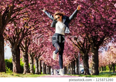 Dynamic scenic young adult pretty happy woman enjoy walking spring city street blooming pink cherry trees alley background in Magdeburg city. Female person joyful carefree portrait smiling outdoors Dynamic scenic young adult pretty happy woman enjoy walking spring city street blooming pink cherry trees alley background in Magdeburg city. Female person joyful carefree portrait smiling outdoors 125871260