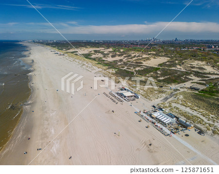 Aerial view of a wide sandy beach with beach bars and dunes, stretching along the coast with a distant city skyline visible under a bright blue sky. 125871651