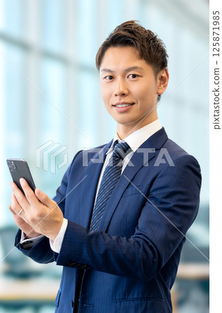拿著智慧型手機的微笑的男性商人 拿著智慧型手機的微笑的男性商人 125871985