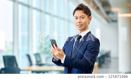 拿著智慧型手機的微笑的男性商人 拿著智慧型手機的微笑的男性商人 125871986