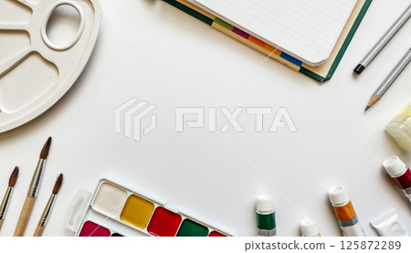 Creative workspace with watercolor paints, paintbrushes, color tubes, sketchbooks, and pencils arranged around a white background 125872289