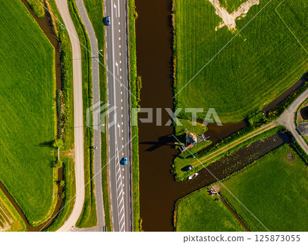 Aerial view of a Dutch countryside with a traditional windmill by a canal, green fields, and a nearby road with moving cars under a clear blue sky. Aerial view of a Dutch countryside with a traditional windmill by a canal, green fields, and a nearby road with moving cars under a clear blue sky. 125873055