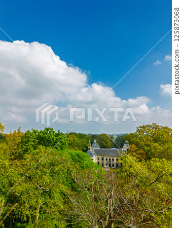 Aerial view of a historic castle surrounded by a forest, with vast green fields, winding waterways, and a river stretching across the flat Dutch countryside under a partly cloudy sky. 125873068