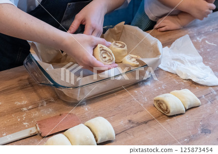 Baking dish with raw cinnamon rolls on table in kitchen and woman's and child's hands Baking dish with raw cinnamon rolls on table in kitchen and woman's and child's hands 125873454