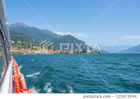 Menaggio nestled by Lake Como showcases colorful buildings and boats gently bobbing in the water, capturing the essence of Italian lakeside charm on a bright sunny day. 125873934