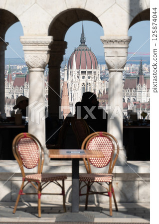 Outdoor cafe terrace with red and white chairs and stone arches framing a view of the Hungarian Parliament Building in Budapest 125874044