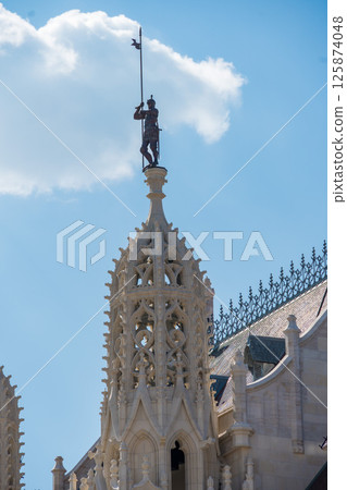 Ornate spire with a statue of a medieval knight holding a flag on the roof of Matthias Church in Budapest 125874048