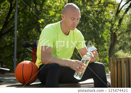 Basketball player eating energy bar or protein bar and drinking water while resting after training at stadium. Healthy protein sugar free snack dieting food for sporty lifestyle. 125874421