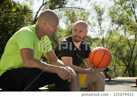 Two basketball players sitting on bench and discussing game on sports court. Sports competition game. Outdoor workout. Team sport. Two basketball players sitting on bench and discussing game on sports court. Sports competition game. Outdoor workout. Team sport. 125874422