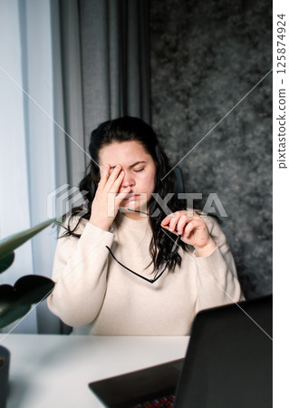 Symptoms of overworking. Young tired female freelancer in eyeglasses sitting in front of computer rubbing eyes feeling eye strain during work 125874924