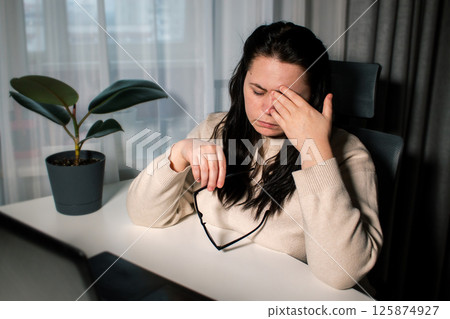 Symptoms of overworking. Young tired female freelancer in eyeglasses sitting in front of computer rubbing eyes feeling eye strain during work 125874927