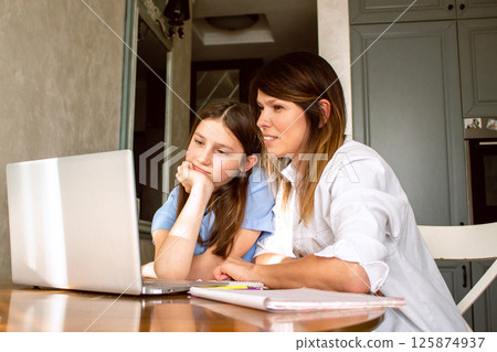 Mother and daughter using laptop sitting together in home kitchen. Mom helping kid with school homework. Education with modern technologies. 125874937
