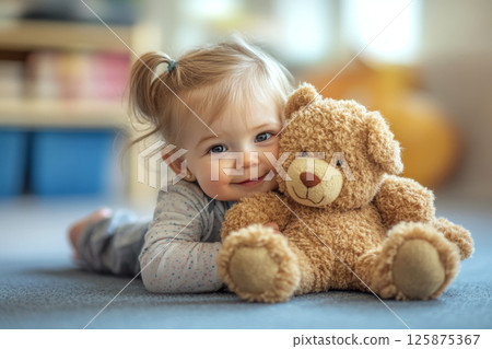 Child lying on a carpet cuddling a soft toy bear in a cozy, blurred background setting. 125875367