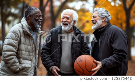 Three elderly men with gray beards laugh together outdoors, one holding a basketball in autumn. Concept of friendship and leisure. For active lifestyle promotion. 125875448