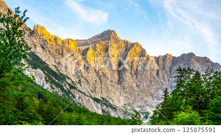 Triglav Mountain stands tall in the Julian Alps, bathed in golden light during sunset. Lush greenery at its base highlights the rugged texture of the rock faces. 125875591