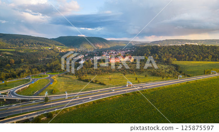 A serene dusk view over Zbraslav, featuring the villages picturesque buildings with rolling hills in the background and a highway in the foreground. A serene dusk view over Zbraslav, featuring the villages picturesque buildings with rolling hills in the background and a highway in the foreground. 125875597