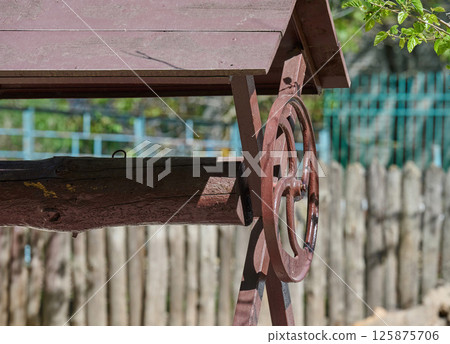 Vintage wooden water well with a hand crank and weathered roof, set against a rustic wooden fence and garden background 125875706