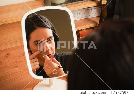 A young girl sits near a small mirror. Attributes for applying makeup next to the girl. In the mirror, a reflection of a girl with short black hair. A young girl sits near a small mirror. Attributes for applying makeup next to the girl. In the mirror, a reflection of a girl with short black hair. 125875921