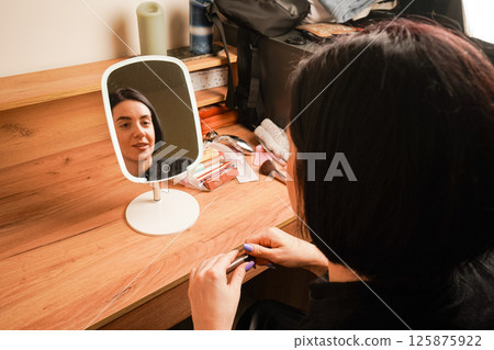 Attributes for applying makeup next to the girl. In the mirror, a reflection of a girl with short black hair. A young girl sits near a small mirror. Attributes for applying makeup next to the girl. In the mirror, a reflection of a girl with short black hair. A young girl sits near a small mirror. 125875922
