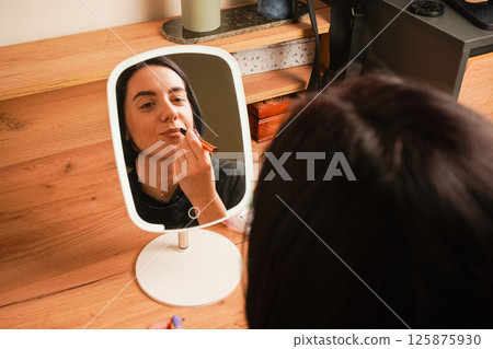 A close-up of a woman applying makeup shows her lining her lips. Her relaxed expression reflects self-care. Behind her, a wooden shelf and small decorative items create a cozy atmosphere. 125875930