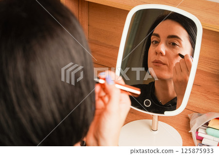 Behind her, a wooden shelf and small decorative items create a cozy atmosphere. A close-up of a woman applying makeup shows her lining her lips. Her relaxed expression reflects self-care. 125875933