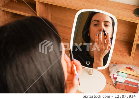 Her relaxed expression reflects self-care. Behind her, a wooden shelf and small decorative items create a cozy atmosphere. A close-up of a woman applying makeup shows her lining her lips. 125875934