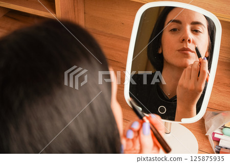 Her short black hair adds elegance to her look. The room is accented by warm wooden tones. Applying mascara to her brows, a woman admires her reflection in a hand-held mirror. Her short black hair adds elegance to her look. The room is accented by warm wooden tones. Applying mascara to her brows, a woman admires her reflection in a hand-held mirror. 125875935