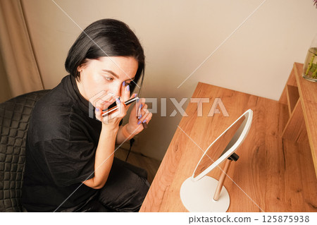 The room is accented by warm wooden tones. Applying mascara to her brows, a woman admires her reflection in a hand-held mirror. Her short black hair adds elegance to her look. 125875938