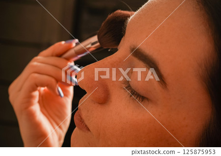Her eyes are closed, highlighting her focus on self-care. Her black bob haircut and natural complexion stand out. A young woman gently applies powder to her face using a makeup brush. Her eyes are closed, highlighting her focus on self-care. Her black bob haircut and natural complexion stand out. A young woman gently applies powder to her face using a makeup brush. 125875953