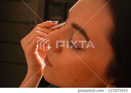 Her eyes are closed, highlighting her focus on self-care. Her black bob haircut and natural complexion stand out. A young woman gently applies powder to her face using a makeup brush. Her eyes are closed, highlighting her focus on self-care. Her black bob haircut and natural complexion stand out. A young woman gently applies powder to her face using a makeup brush. 125875955