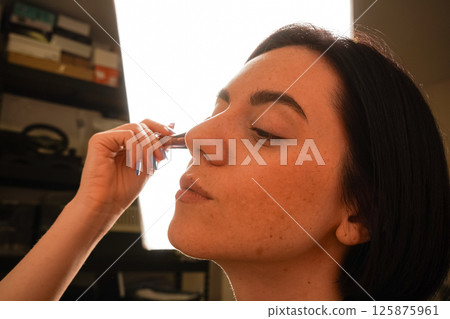 Her eyes are closed, highlighting her focus on self-care. Her black bob haircut and natural complexion stand out. A young woman gently applies powder to her face using a makeup brush. Her eyes are closed, highlighting her focus on self-care. Her black bob haircut and natural complexion stand out. A young woman gently applies powder to her face using a makeup brush. 125875961