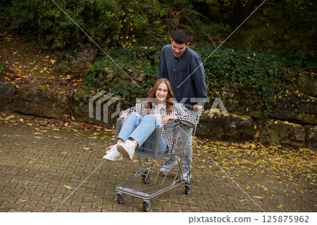 Happy young couple having fun with a shopping cart outdoors in autumn park, wearing stylish casual outfits with denim and flannel, laughing and enjoying the moment. 125875962