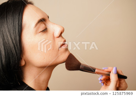 The warm lighting complements her natural skin tone. Her dark hair and serene expression create a calm atmosphere. Applying makeup with a soft brush, a woman focuses on perfecting her look. 125875969