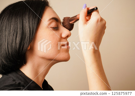 Her dark hair and serene expression create a calm atmosphere. Applying makeup with a soft brush, a woman focuses on perfecting her look. The warm lighting complements her natural skin tone. Her dark hair and serene expression create a calm atmosphere. Applying makeup with a soft brush, a woman focuses on perfecting her look. The warm lighting complements her natural skin tone. 125875977
