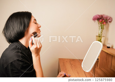 Her natural beauty and black hair shine in the warm light. The woman sits at a makeup table with a small mirror in front of her. A woman carefully applies powder to her face with a large makeup brush. 125875980